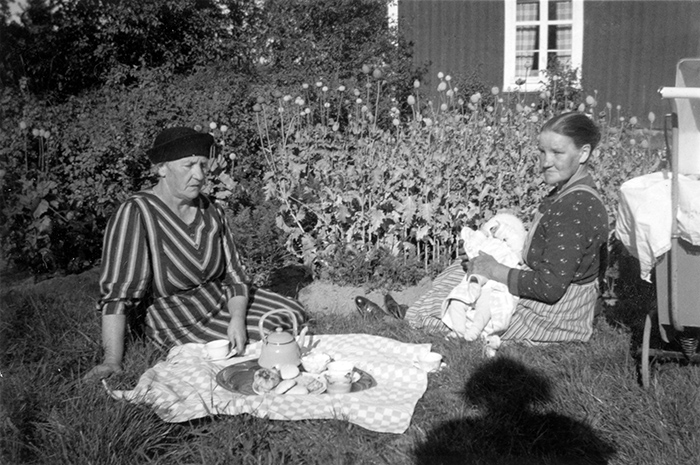 Två kvinnor sitter på gräsmatta med filt, kaffekanna och kakfat. Kvinnan till vänster har ett spädbarn i famnen.