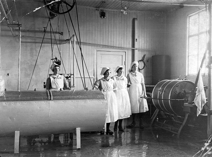 Interiörbild från mejeri med blanka golv. Fyra kvinnor med vita förkläden och mössor står vid stora cylinderformade behållare.