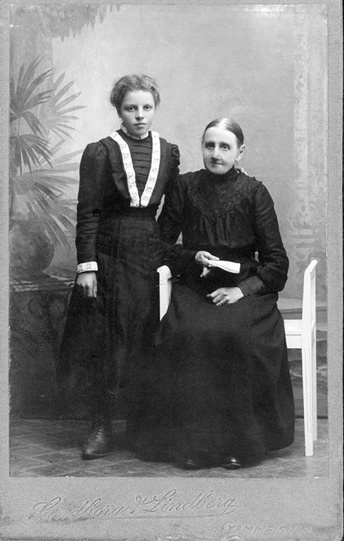 En ung flicka står bredvid en gammal kvinna sittande i stol. Båda i svart klänning.