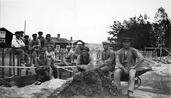 Gruppbild av män som arbetar vid bygge. Två sitter på en skottkärra. I förgrunden en jordhög, i bakgrunden syns några mindre trähus.