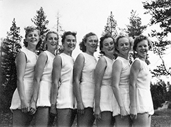 Sju unga kvinnor på rad i vita gymnastikkläder.