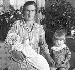 Kvinna sittande med spädbarn i famnen och liten dotter på stol bredvid.