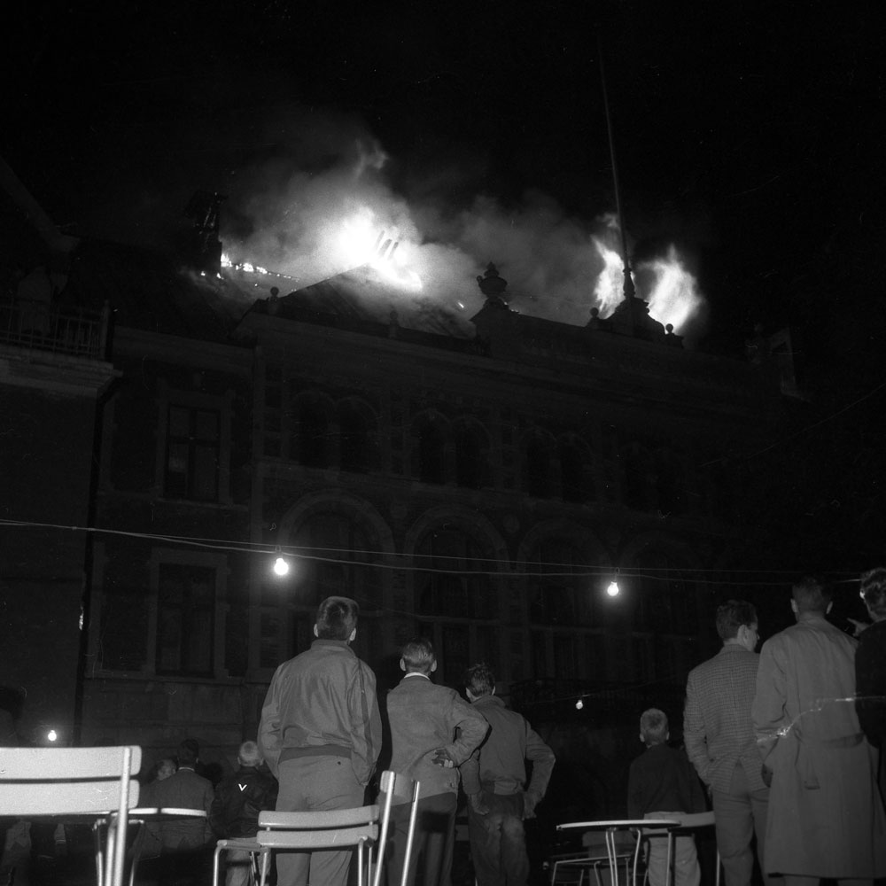 Åskådare ser på när stadshotellet brinner. Mörkt förutom elden på taket.