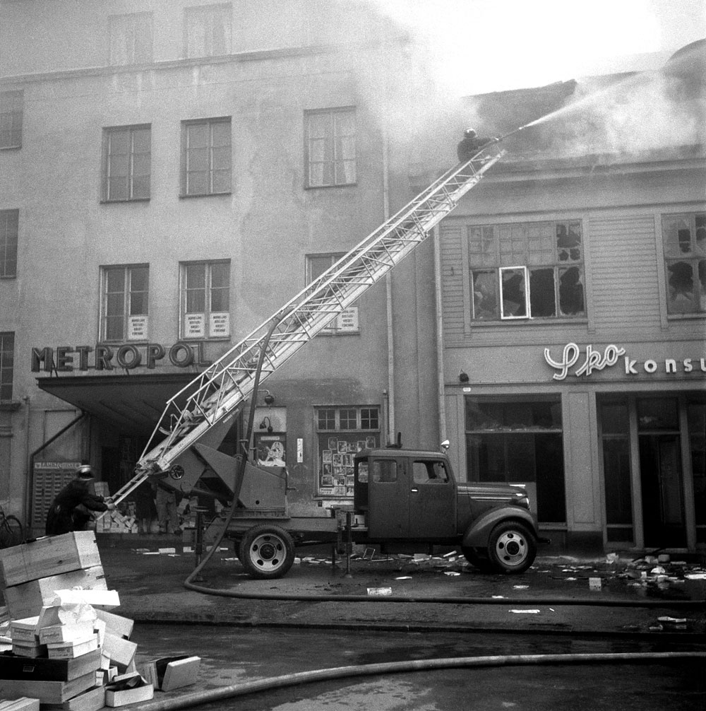 Brandbil med stege vid hus i staden, brandman sprutar vatten stående högst upp på stegen.
