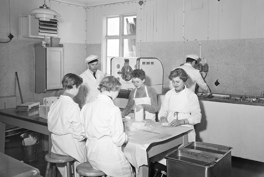 Vitklädda kvinnor sitter och slår in glasspinnar en och en för hand.  Man i vit keps tittar på och en annan man arbetar vid en bänk. 