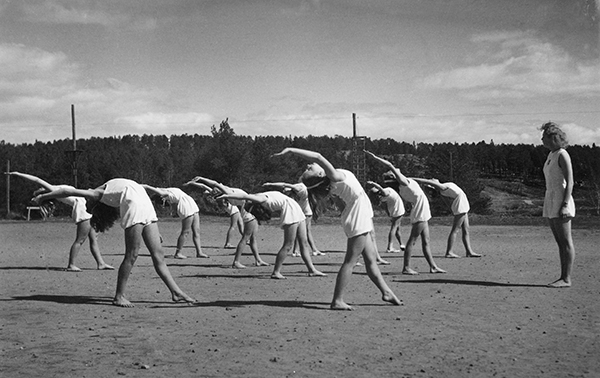 Kvinnliga gymnaster med bakåtböjda kroppar stående på grusplan utomhus.