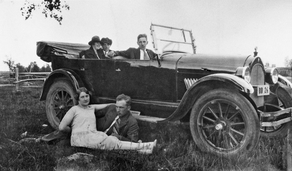 Bil av äldre modell. I bilen sitter tre personer och framför den ligger en man och en kvinna i gräset.