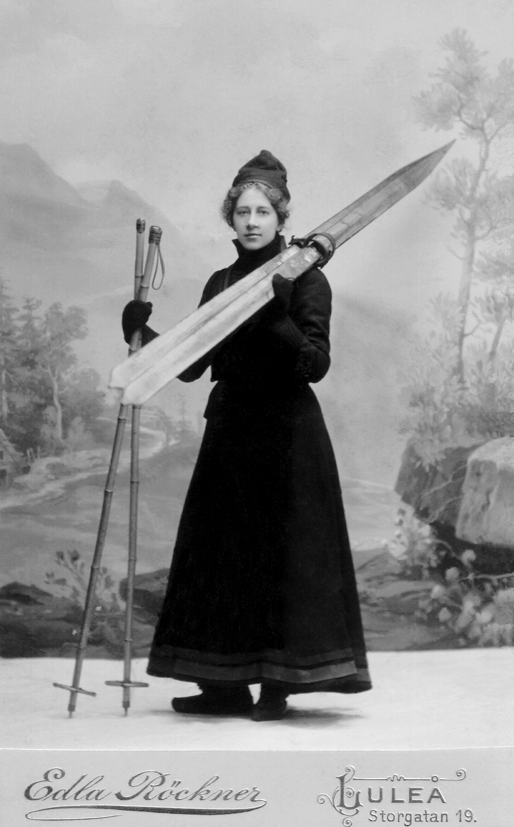 Studiofoto av kvinna i svart långklänning med skidor på ena axeln och skidstavar i handen.