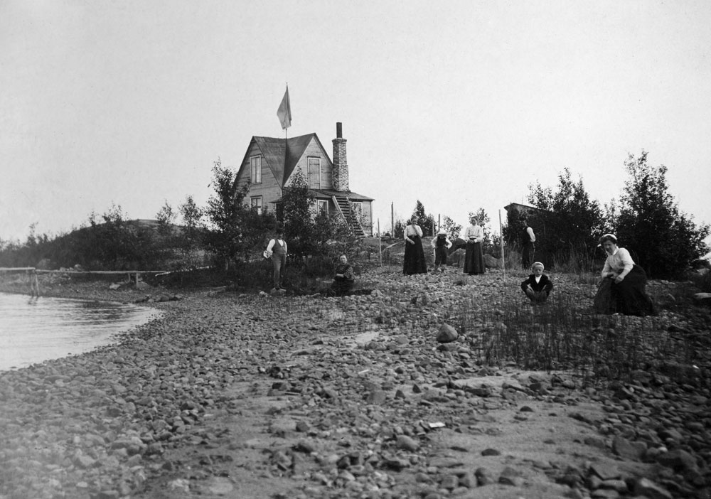 Kvinnor i mörka långklänningar på stenig strand, trähus i bakgrunden.