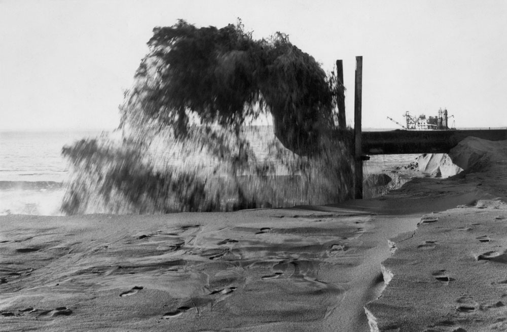 Bild från sandstrand, vid maskin sprutar det upp sand i luften.