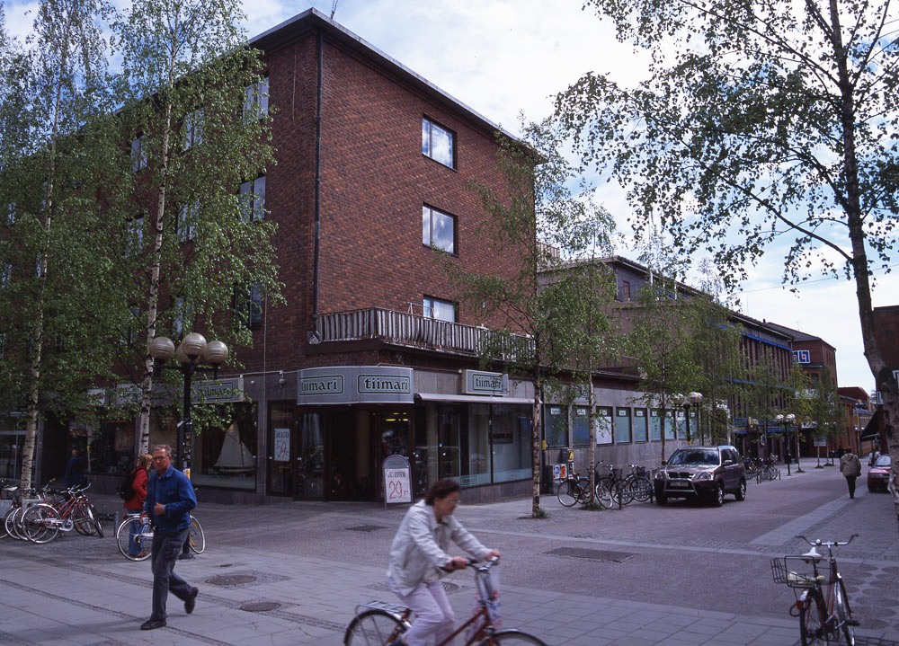 Gatukorsning med butikslokaler i brun tegelbyggnad. Gående och cyklande personer på gatan framför.