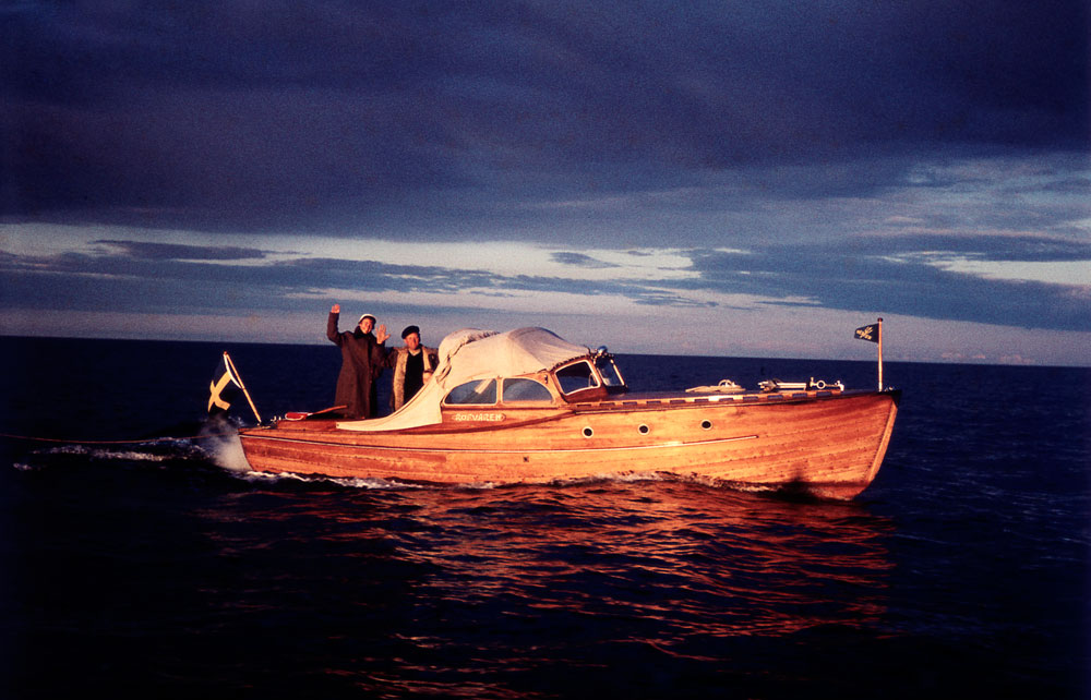 Två män vinkar på mindre båt i kvällsljus.