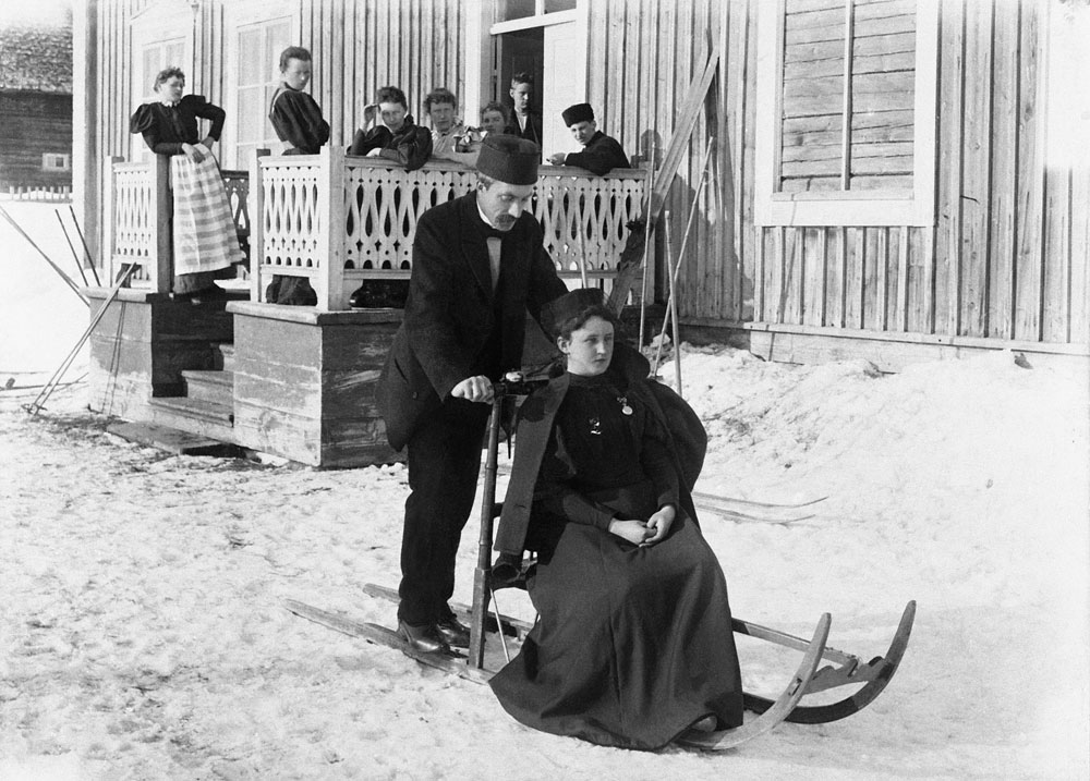 Kvinna i svart långklänning sitter på spark som styrs av man i kostym. Betraktas i bakgrunden av flera personer på farstukvist.