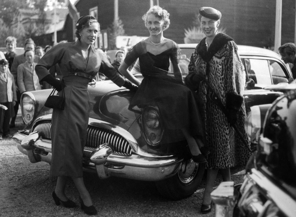 Tre kvinnor i vackra klänningar och hattar sittande på blank amerikansk bil.