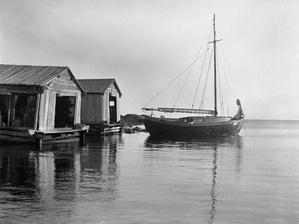 Segelbåt vid sjöbodar av trä, lugnt vatten.