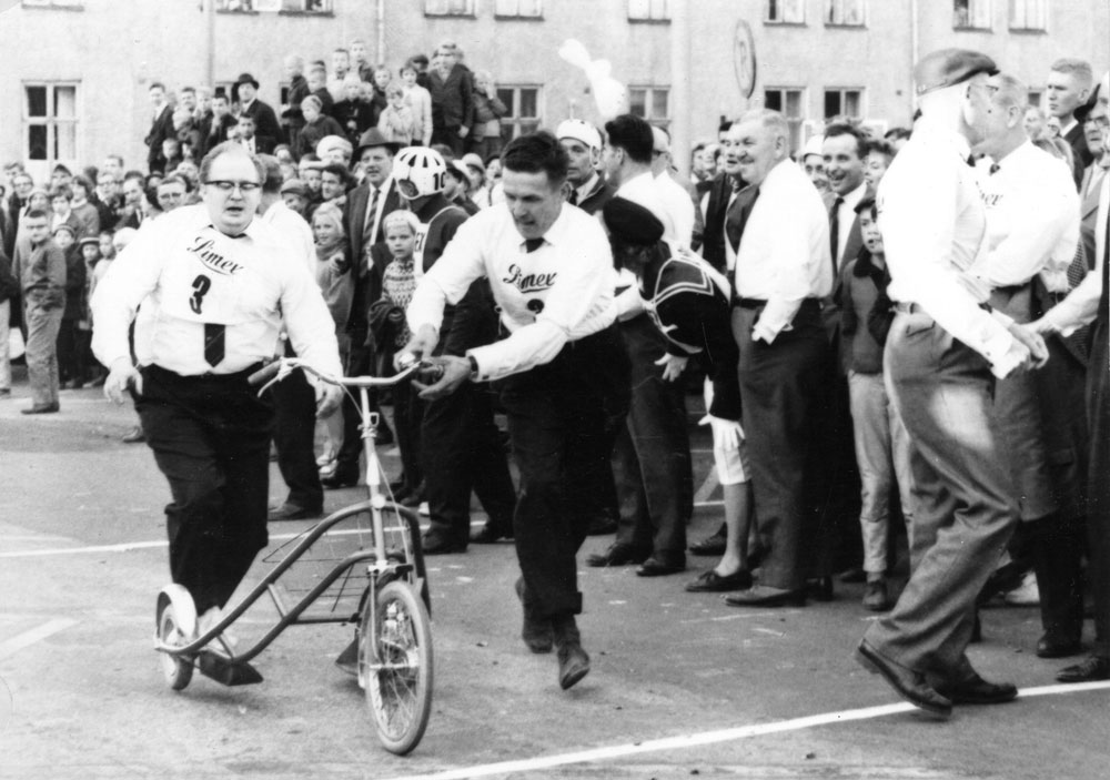 Två män i vit skjorta och mörka byxor springer bakom sparkcykel, folkskara ser på.