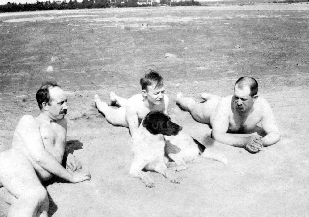 Tre nakna män ligger på strand med en hund.