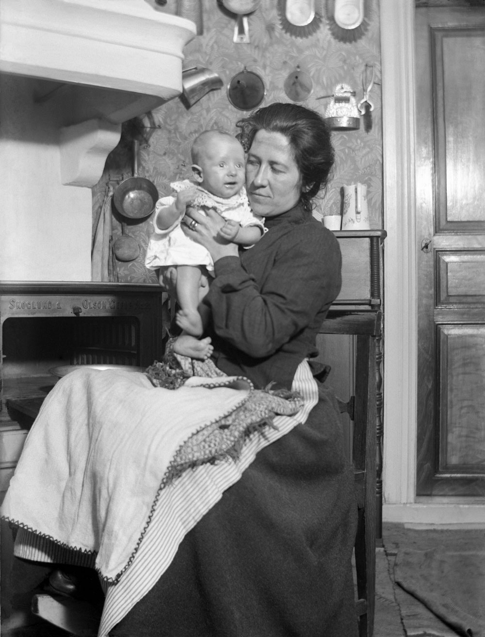 Kvinna med mörk klänning och förkläde sitter vid gjutgärnsspis och håller i ett spädbarn.