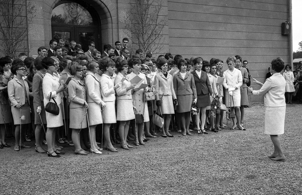Gruppbild av unga kvinnor och män i fina kläder. Kvinna står framför och dirigerar.