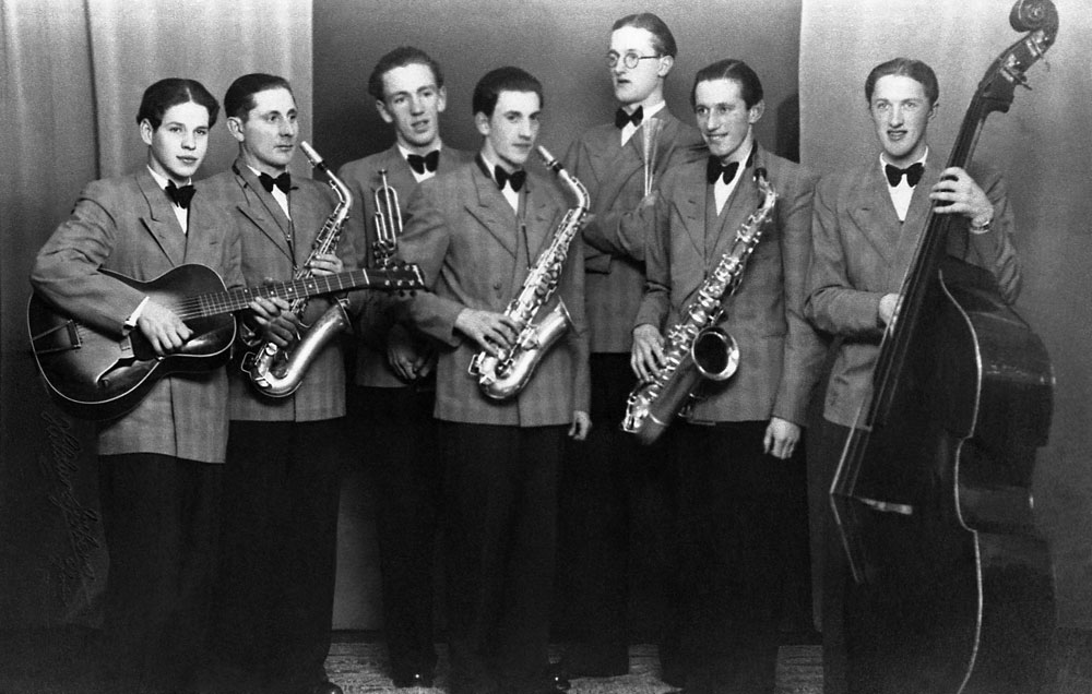 Orkester med sju män i likadana kostymer och fluga spelande gitarr, saxofon, trumpet och basfiol.