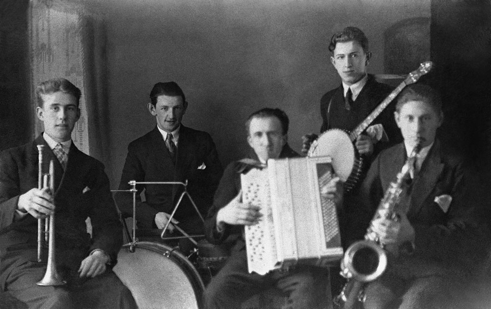 Orkester med fem män som spelar trumpet, trummor, dragspel, banjo och saxofon.