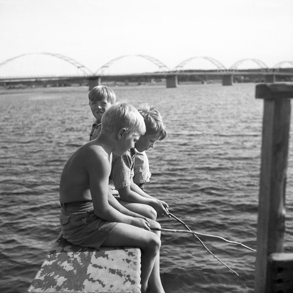 Pojkar sitter och metar vid vatten. Bro i bakgrunden.