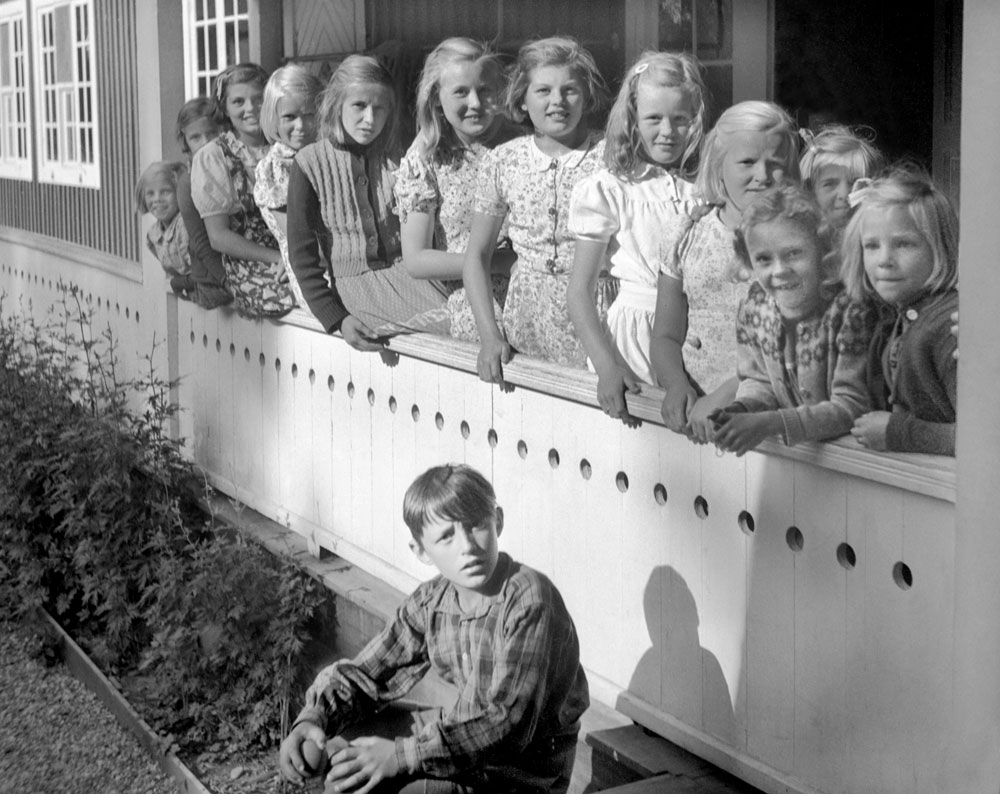 Rad av flickor sittande på verandastaket, nedanför en pojke.