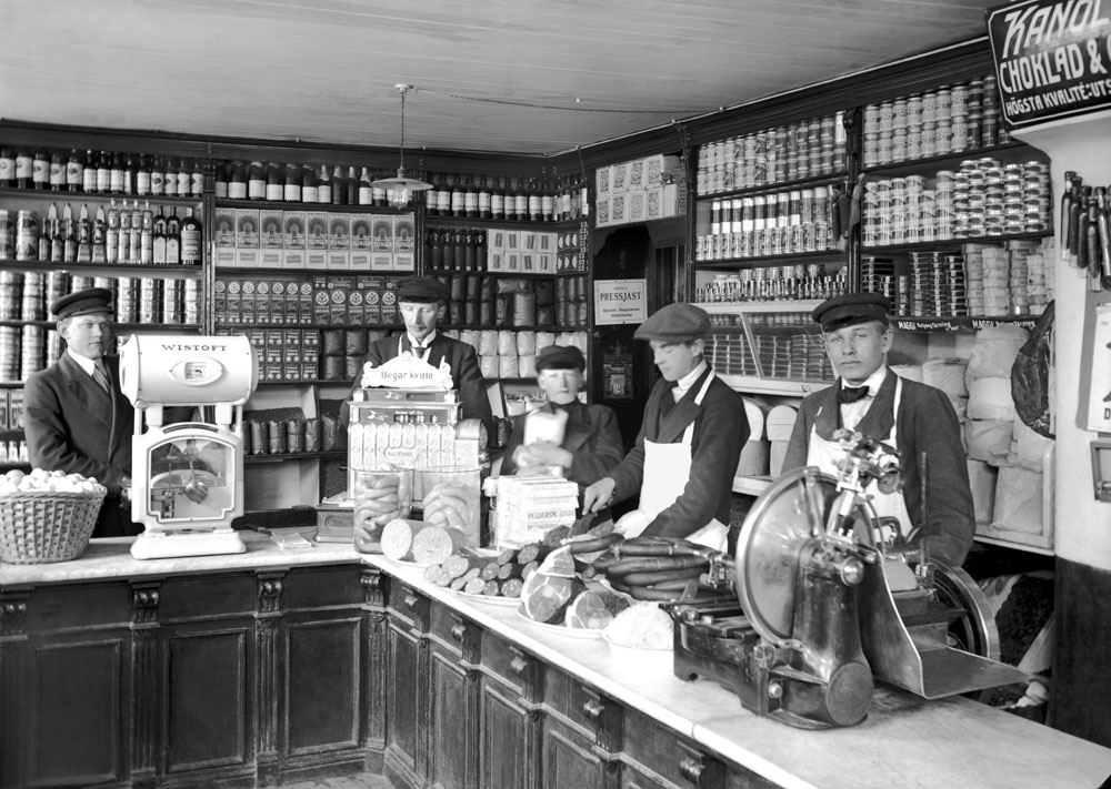 Interiör från livsmedelsbutik 1915 med personal stående bakom disken.