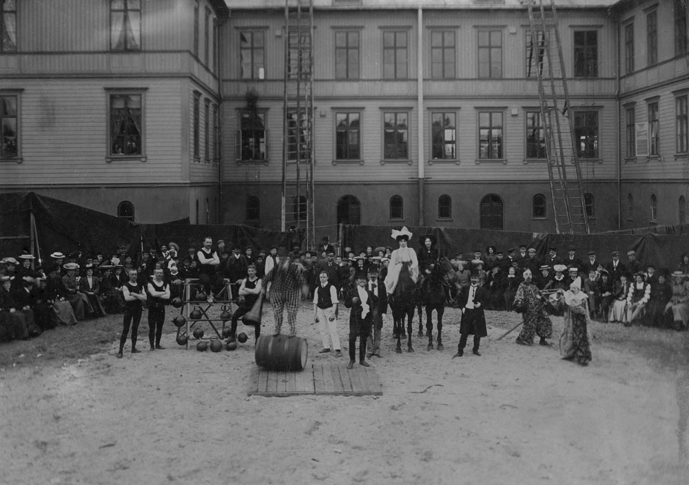 Cirkusartister uppträder för publik framför stor skolbyggnad av trä.