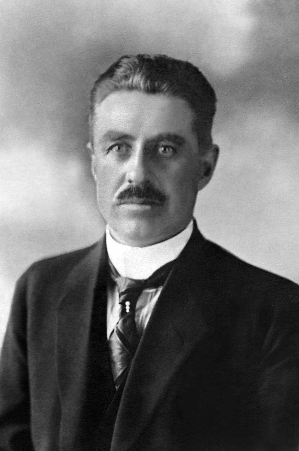 Porträtt i halvfigur av man med mustasch i mörk kavaj och slips.