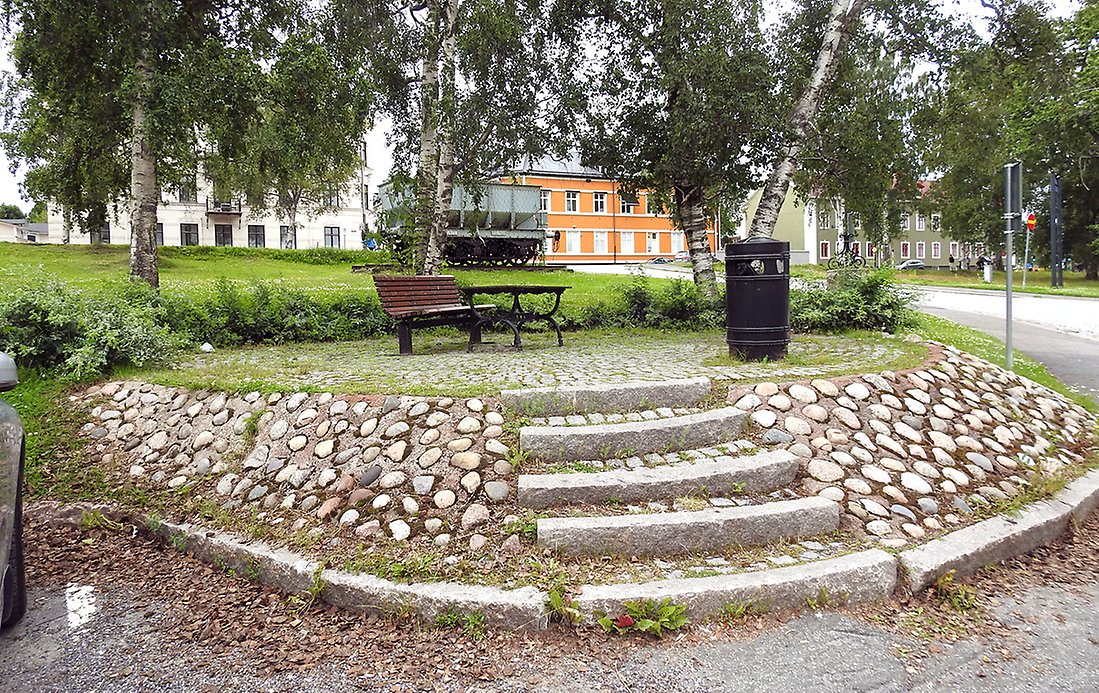 En upphöjd sittplats med trappsteg i sten och stenlagda kanter. Sittplatsen har en parkbänk och ett bord. I bakgrunden syns den gamla järnvägsvagnen